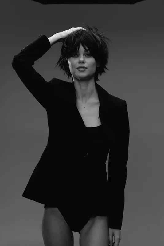 Black and white portrait of a confident woman with short hair wearing a black jacket, posing with her hand on her head.