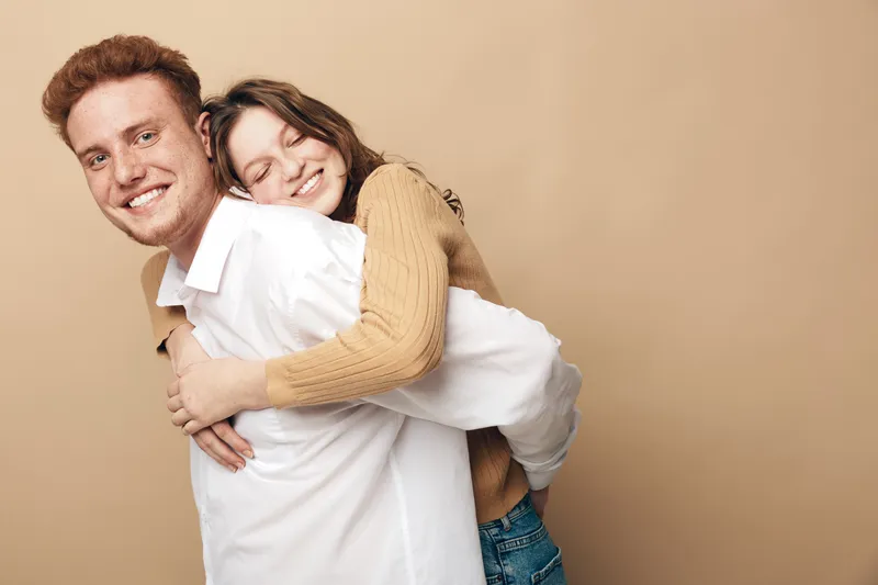 Happy young couple with red hair and brown hair smiling and hugging each other on beige background. Concept of love, friendship, affection, and casual lifestyle together.