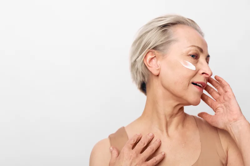 Mature woman with short hair applies skin cream on face with soft touch. Light neutral background highlights natural beauty and skincare routine for aging skin care concept.