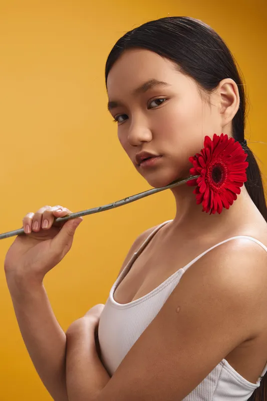 Asian woman in a simple white tank top stands against a vibrant yellow backdrop, holding a red flower near her shoulder and gazing softly at the camera.