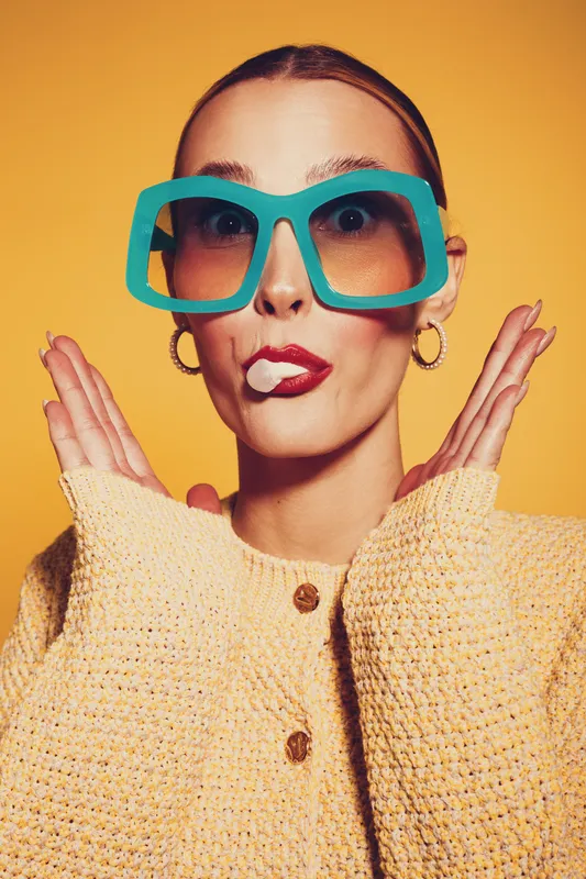 Young woman with oversized turquoise glasses and yellow sweater makes funny face with lipstick on lips against yellow background. Concept of humor, fashion, and playful expression.