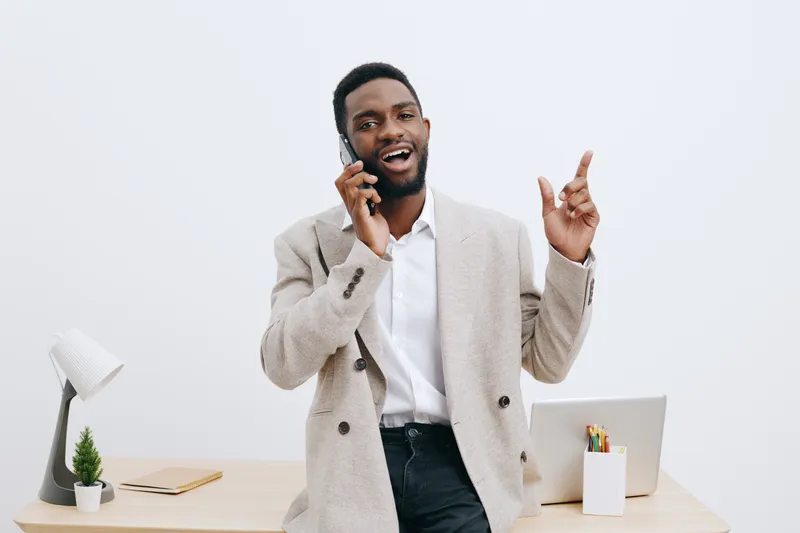 Smiling man talking on mobile phone in modern office workspace with laptop and decor, conveying communication and professionalism, casual business attire, positive mood, light background, studio setti