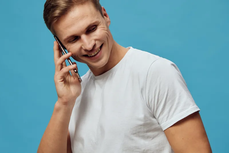 Smiling man talking on mobile phone isolated on plain blue background in casual white t-shirt, cheerful expression, lifestyle concept, communication, technology, happiness, leisure, modern style.