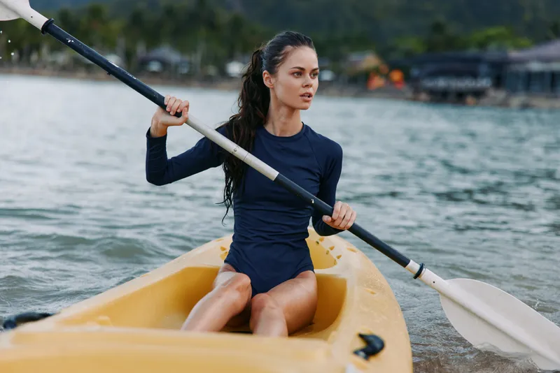 Female paddler sits in a yellow kayak on calm water, wearing a dark athletic suit, gripping a double blade paddle as she concentrates on the stroke.