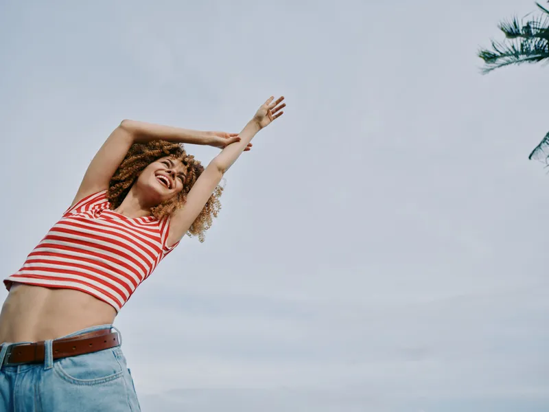 Young woman with curly hair in a red and white striped crop top and light denim jeans stands outdoors, arms raised, grinning, under a clear blue sky, radiating joy.