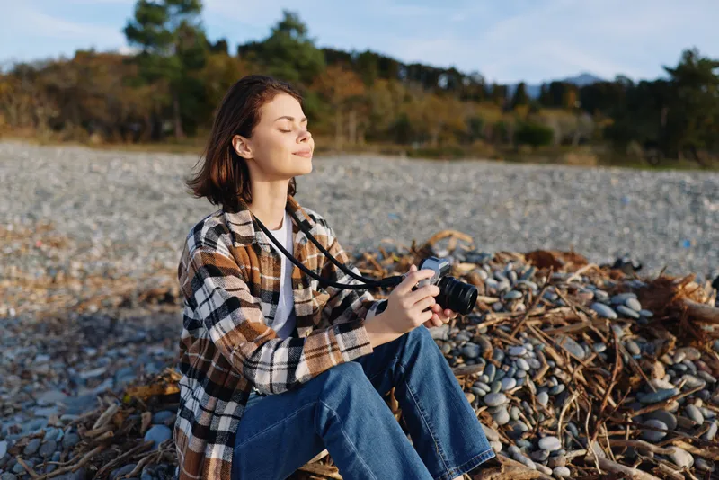 Photographer woman camera outdoor beach rocks nature casual scene with calm expression under natural light on a rocky shore, capturing a peaceful seascape and candid mood