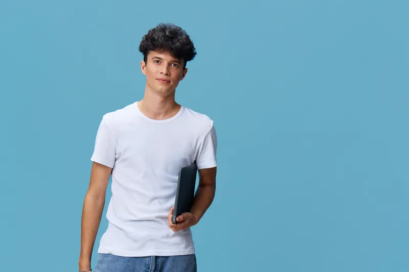 young man holding smartphone wearing casual white t-shirt and jeans standing against plain blue background with relaxed expression and natural hairstyle