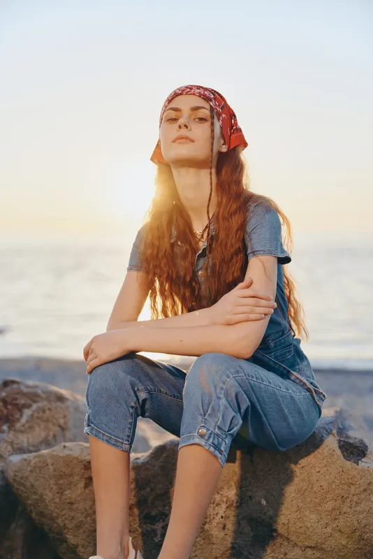 lifestyle woman wearing modern boho-western denim outfit with red bandana sitting outdoors on rocks during golden hour, film color mood, casual street style vibe