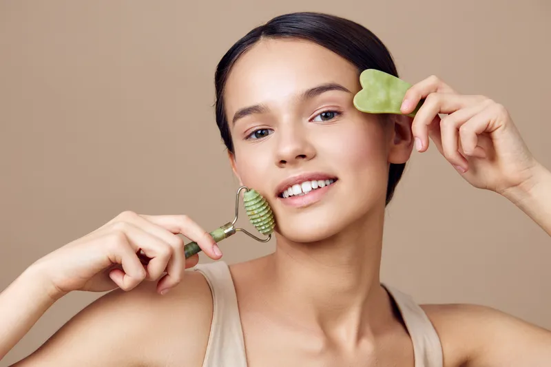 Young woman with a jade roller and gua sha tool, glowing skin, natural beauty, healthy skincare routine, joyful expression, beige background