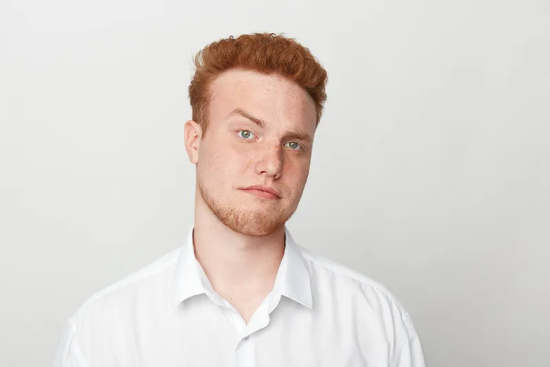 Redhead man with curly hair and beard wears white shirt, looking at camera with neutral expression on light background. Concept of casual style, portrait, and natural look.