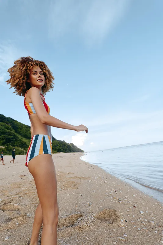 Confident woman on a sandy beach, wearing a colorful bikini, with blue sea and sky, warm sunlight, gentle breeze, and relaxed posture capturing a vibrant summer moment outdoors