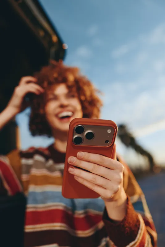 Woman lifestyle, smartphone selfie outdoors, smiling woman with curly hair holds red smartphone, casual striped sweater, shallow depth field, summer sky background, candid moment