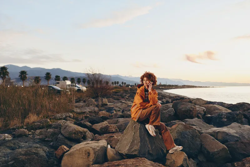 Woman lifestyle on seaside rocks at warm sunset, holding backpack while walking along the shoreline, relaxed travel mood, golden light, distant hills, outdoors nature background