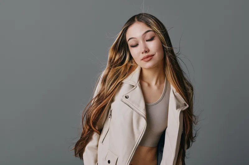 Woman with long hair and closed eyes wearing beige jacket and crop top standing isolated on gray background, casual fashion portrait with calm expression and hairstyle.