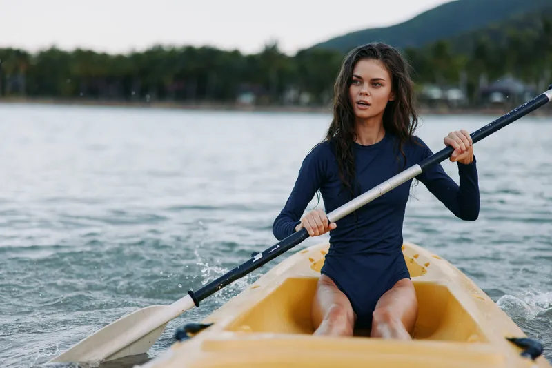 Young female kayaker sits in a yellow kayak, paddling on calm water. She wears a dark wetsuit, focused expression, and moves with strength and balance through the current.