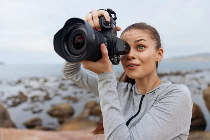 Young woman photographer taking pictures at the seaside, showcasing excitement and creativity with a digital camera, wearing comfortable outdoor clothing
