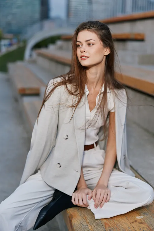 Stylish woman in beige suit sitting outdoors on wooden steps with thoughtful expression, natural light, urban background, fashion concept, autumn season
