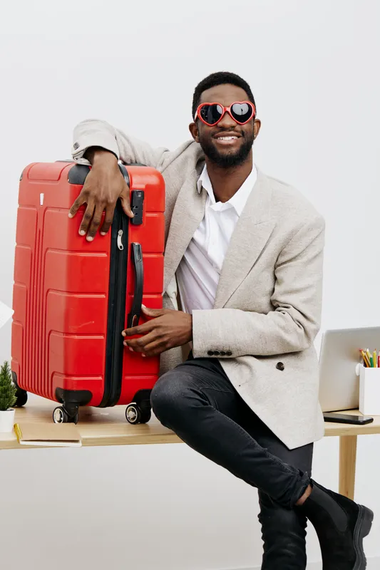Smiling man with dark skin wearing stylish sunglasses and casual beige jacket, holding a large red suitcase, in modern indoor setting. Concept: travel, vacation, holiday, airport, leisure, lifestyle