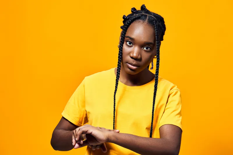 A young Black woman with braided hair wearing a bright yellow shirt, looking thoughtfully at her wrist, set against a vibrant yellow background