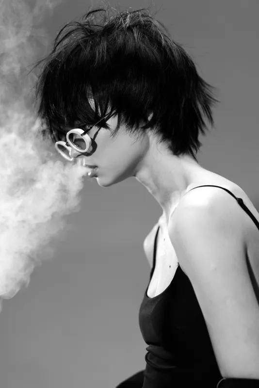 Black and white portrait of a young woman with short hair and sunglasses exhaling smoke in a stylish tank top.