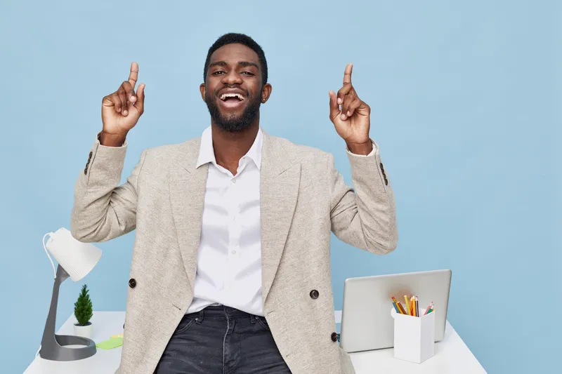 Happy joyful man with dark skin wearing beige blazer and white shirt, raising both hands with fingers pointing up, standing in modern office with desk, lamp, and supplies on blue background. People li