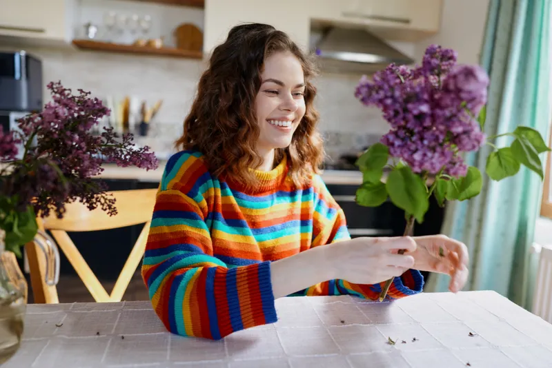 Smiling woman with curly hair wearing a colorful striped sweater sits at a white table at home, using a tablet. Bright flowers and soft light create a warm, cozy lifestyle scene.