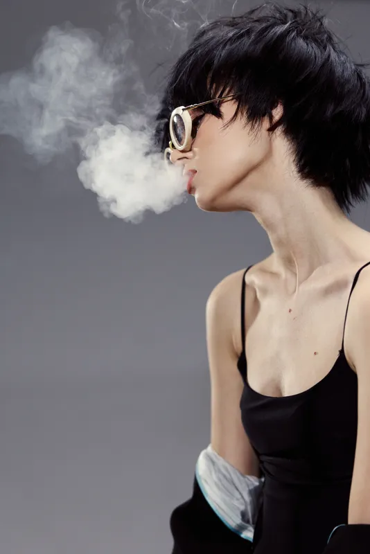 Young woman exhaling smoke wearing stylish glasses and black dress, modern fashion, expressive mood, studio background