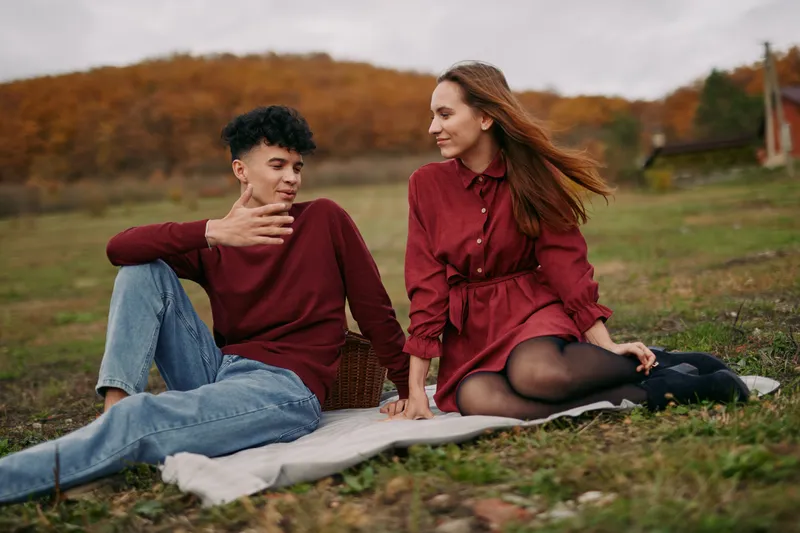 A young couple sits on a blanket in an autumn field, sharing a candid conversation and warm smiles, captured with authenticity and veracity to reflect true everyday connection.