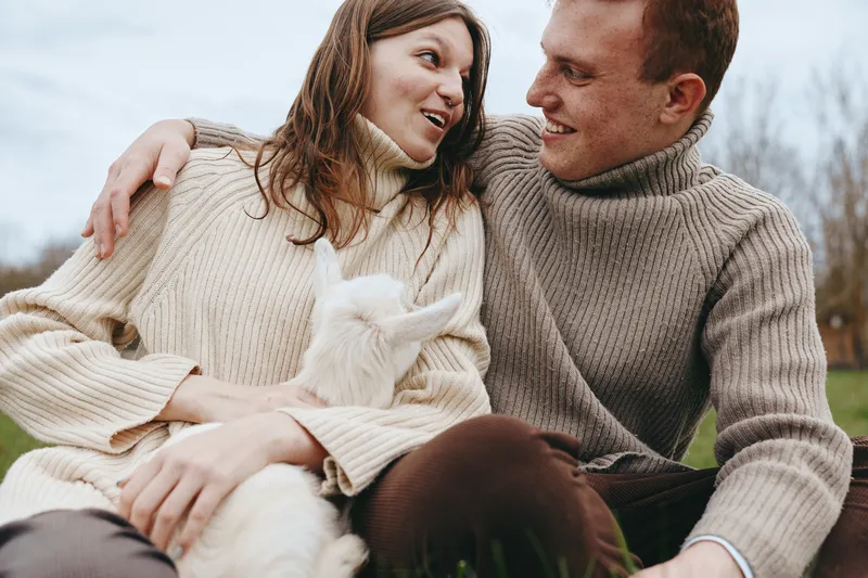 Cheerful couple outdoors, cuddling with a playful goat kid, cozy sweaters, sunlit moment, natural textures, grass beneath, candid lifestyle portrait, warm connection, serene countryside