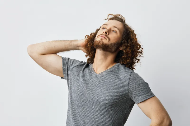 Confident young man with curly hair in casual gray t shirt showcasing self assurance and style against a neutral background