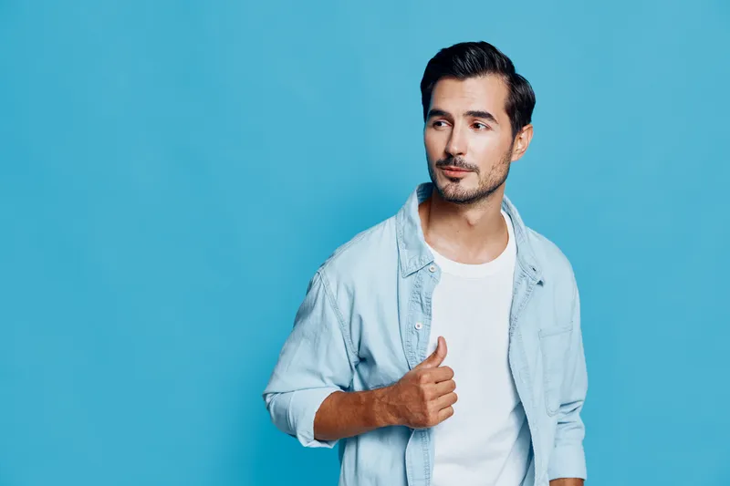 Stylish young man wearing light blue shirt looks confidently at the camera while posing against a solid blue background, showcasing a trendy and modern lifestyle.