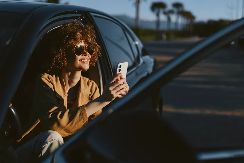 A woman in a car smiles while looking at her phone, wearing sunglasses and casual clothing. The retro film color tone adds warmth to a relaxed lifestyle moment.