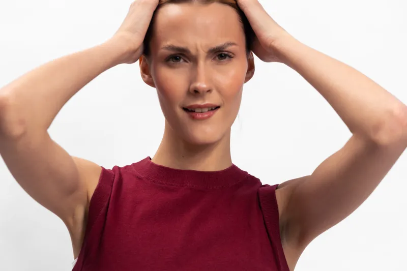 A frustrated young woman in a sleeveless red top holds her head with both hands, showing tension and stress on a white background. Concept for anxiety, pressure, and emotional struggle.