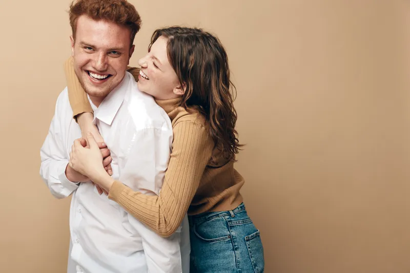 Happy young couple in casual clothes smiling and hugging each other on beige background. Concept of love, friendship, relationship, and positive emotions.