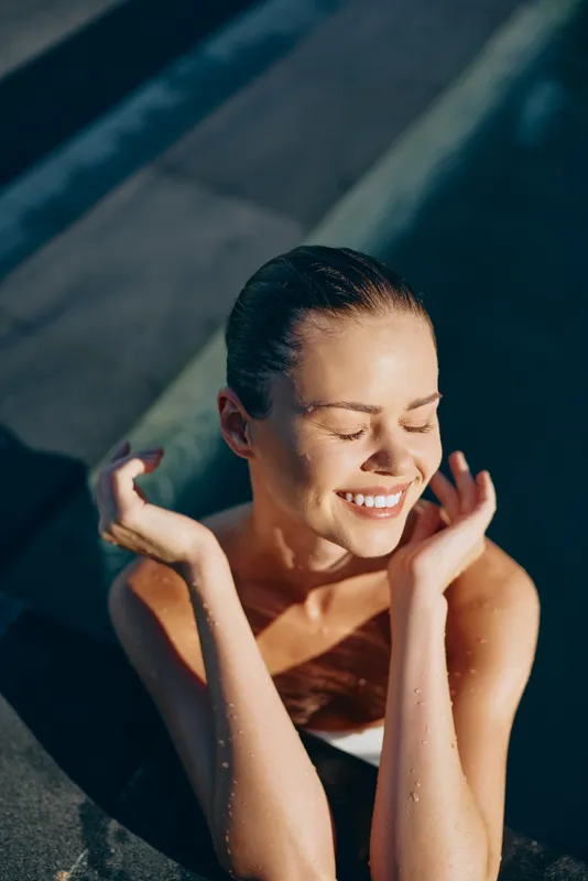 Lifestyle portrait of a joyful woman by a pool, cooling off under sun, eyes closed, warm smile, relaxed moment, natural candid mood, serene summer scene by water