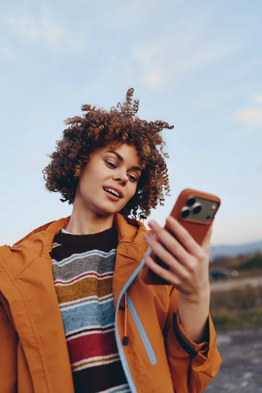 Woman with curly hair takes a selfie outdoors, holding a phone and smiling in an orange jacket over a striped sweater, mountain landscape at sunset, lifestyle moment, soft natural light