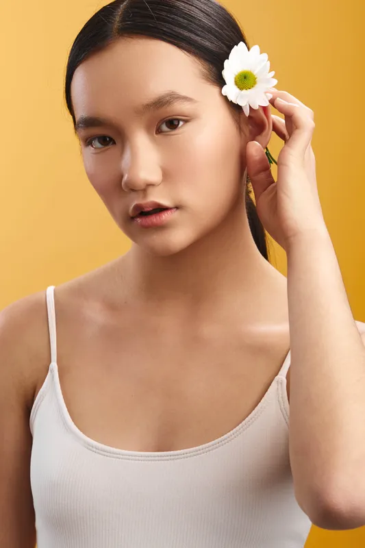 asian woman poses in a bright studio, wearing a white tank top and a flower in her hair, exuding calm confidence and natural beauty against a clean yellow backdrop.
