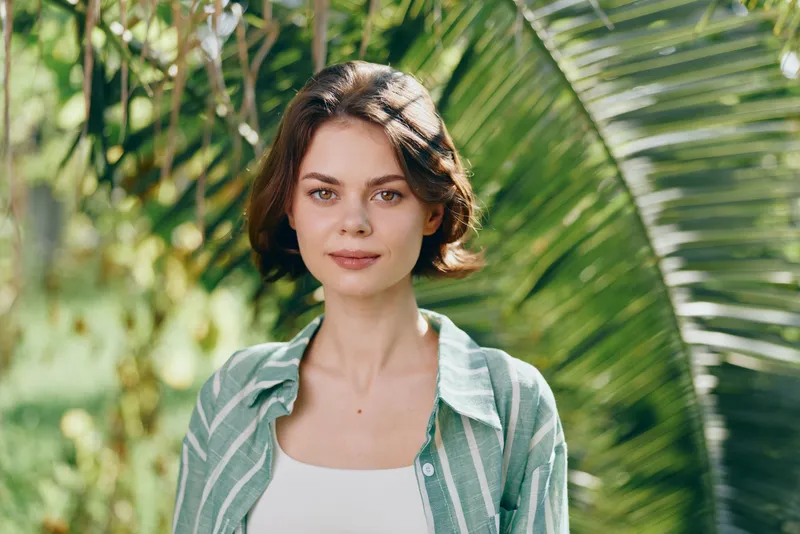 Portrait of a young woman outdoors with lush greenery, soft lighting, and a calm, attentive expression.
