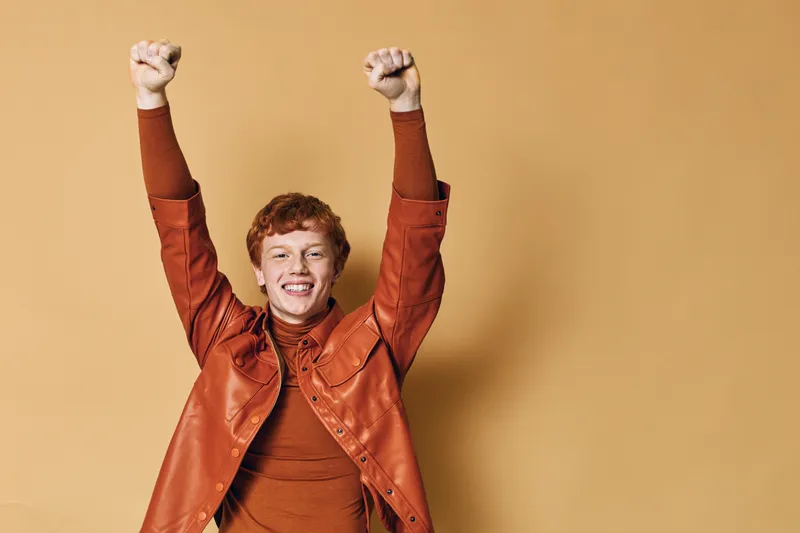 Young man with red hair wearing brown jacket and sweater raising hands in excitement smiling on beige background in studio. Happy male celebrating success or achievement indoors.