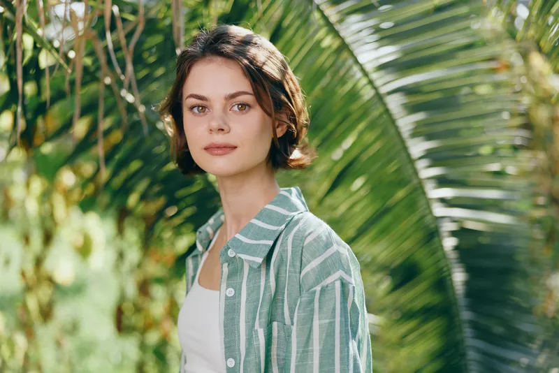 Woman in a striped shirt stands in a sunlit garden, soft natural light highlighting her thoughtful expression. Lush greenery surrounds her, creating a serene portrait that conveys calm, confidence,