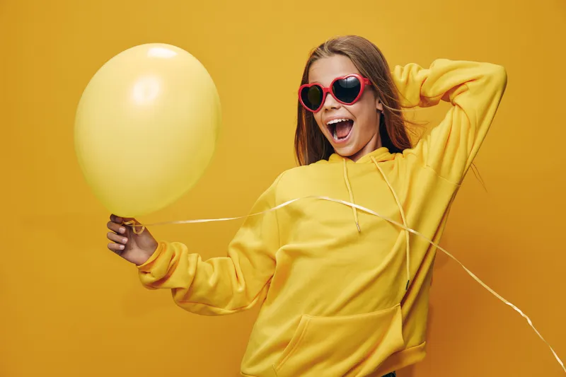 Young woman in yellow hoodie and heart shaped sunglasses holding yellow balloon with excited expression on yellow background for celebration and fun.