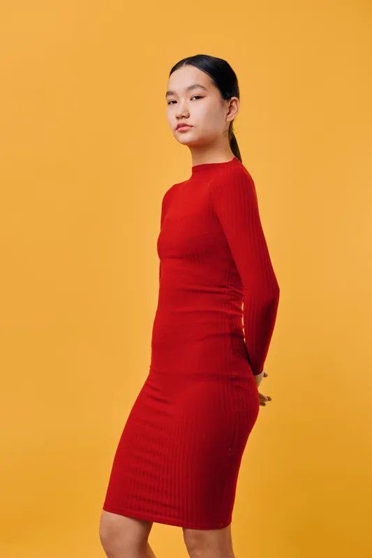 Young woman posing in red fitted dress against solid yellow background, standing with hands behind back and confident facial expression in studio portrait.