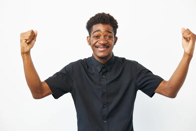 Happy black man celebrating success with raised fists, showcasing joy and enthusiasm in a sleek, modern attire against a clean, white background.