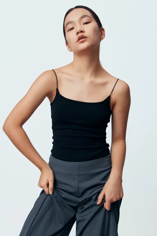 Young woman posing in black tank top and gray pants on white background. Fashion model with hands on hips looking confident and stylish in studio portrait image.