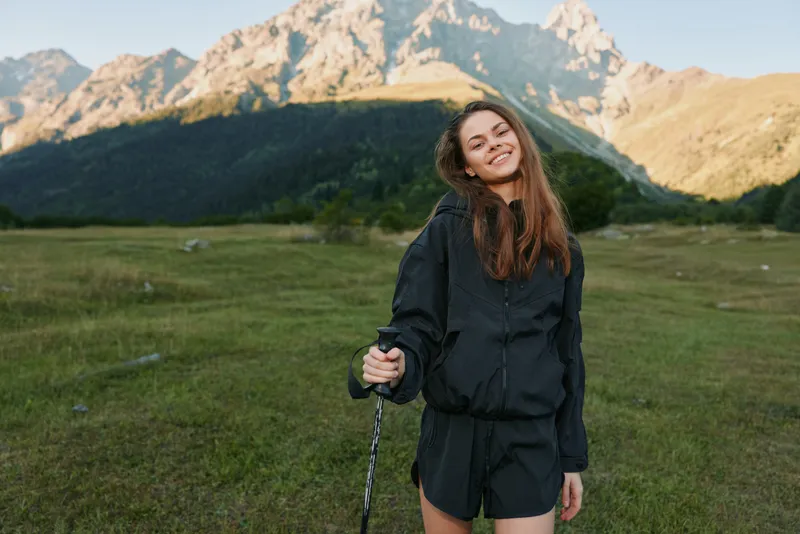 A cheerful young woman stands on a grassy meadow with towering mountains in the background. She smiles, wearing hiking gear, and holds a trekking pole, enjoying the outdoors.