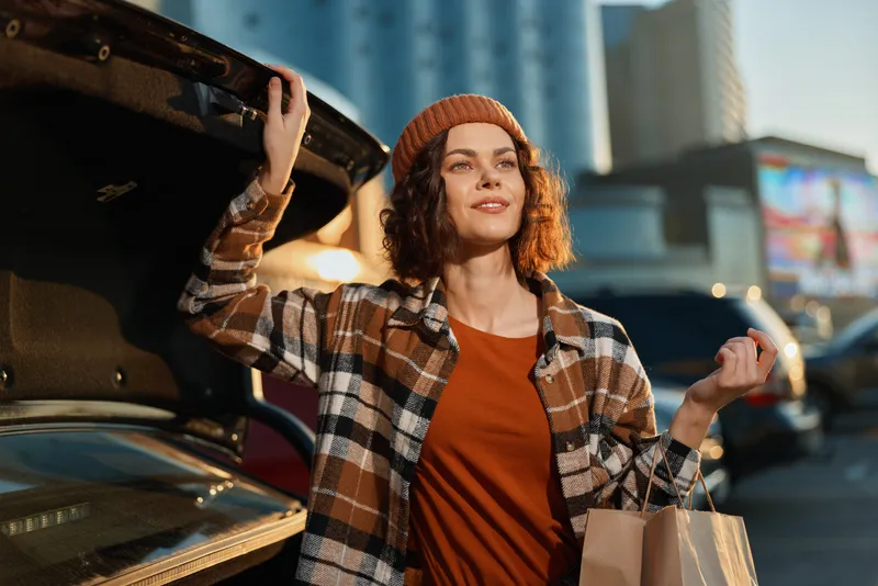 Woman shopping by car trunk in city street at golden hour, sunlight on face creating golden hour glow; candid lifestyle portrait conveying authenticity, mindful living and emotional storytelling.