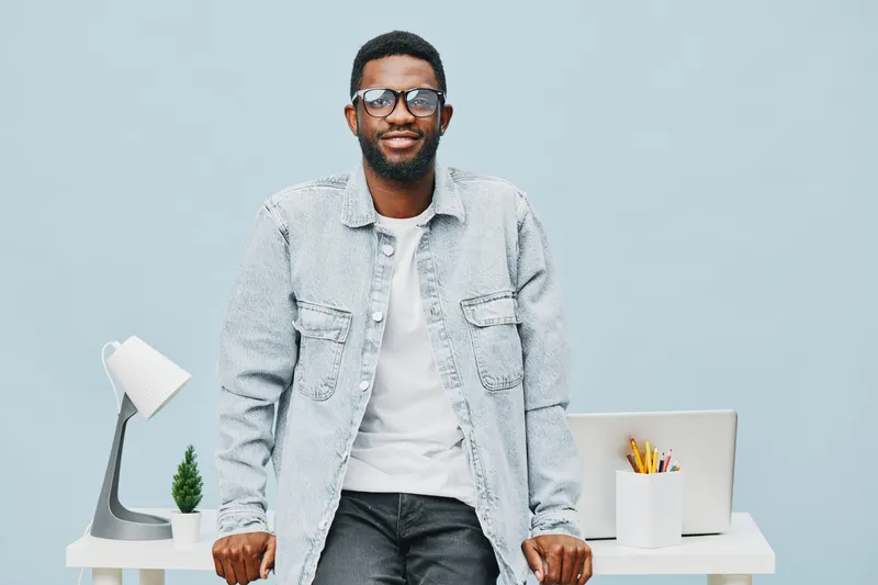 Confident young man with glasses and a light denim jacket sitting in modern office environment smiling, casual clothing, bright background, interior decor, lifestyle concept, positive mood, contempora