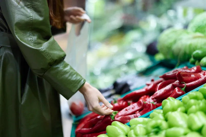 Женщина в продуктовом магазине выбирает свежие овощи для приготовления ужина дома, держа в руках красный перец на рынке