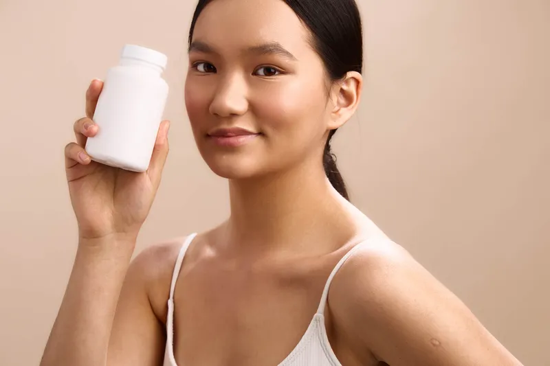 asian woman holds a white bottle in a clean studio, presenting skincare product with calm confidence. she gazes softly at the camera, signaling trust and a healthy routine in a modern beauty setting.