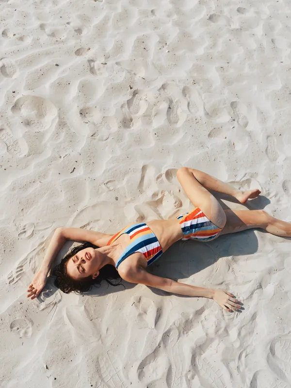 A relaxed woman lies on sunlit white sand wearing a striped bikini, creating a casual beach vibe with soft shadows, bright light, and a carefree pose.